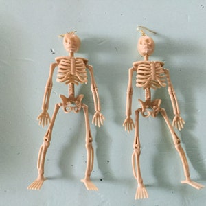 May include: Two beige plastic skeleton earrings with gold-colored ear wires. The earrings are in the shape of a human skeleton, with articulated limbs and a skull. The earrings are displayed against a light blue background.
