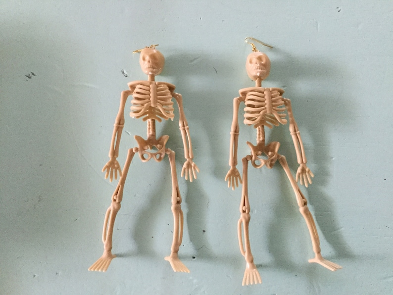 May include: Two beige plastic skeleton earrings with gold-colored ear wires. The earrings are in the shape of a human skeleton, with articulated limbs and a skull. The earrings are displayed against a light blue background.
