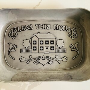 May include: A rectangular, pewter-colored tray with rounded corners. The center features an embossed design with the words "BLESS THIS HOUSE" above a house, trees, and floral accents. A heart is in the center of the house.
