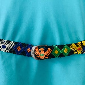 May include: A beaded belt with a silver buckle. The belt is black with colorful geometric and bird designs in red, white, blue, orange, and yellow. The belt is worn over a turquoise top.