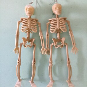 May include: A pair of beige plastic skeleton earrings. Each earring features a full skeleton with articulated limbs and a skull head. The earrings hang from silver-colored fish hook ear wires. Perfect for Halloween or a unique accessory.