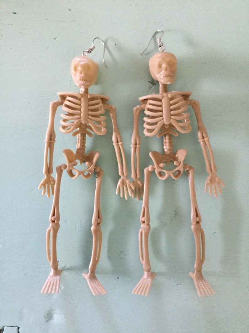 May include: A pair of beige plastic skeleton earrings. Each earring features a full skeleton with articulated limbs and a skull head. The earrings hang from silver-colored fish hook ear wires. Perfect for Halloween or a unique accessory.