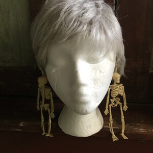 May include: A pair of beige skeleton earrings hanging from gold-colored hooks. The earrings are displayed on a white mannequin head with gray hair. The earrings are a fun, spooky accessory.