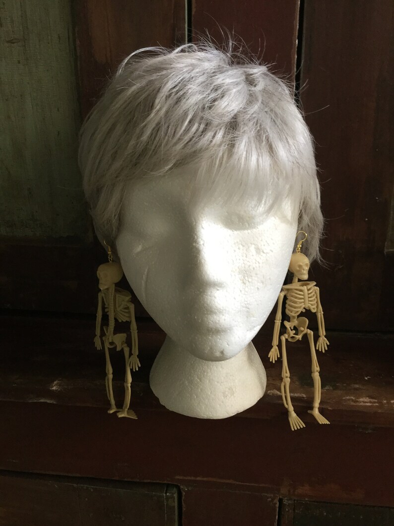 May include: A pair of beige skeleton earrings hanging from gold-colored hooks. The earrings are displayed on a white mannequin head with gray hair. The earrings are a fun, spooky accessory.