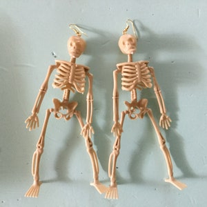 May include: A pair of beige plastic skeleton earrings. Each earring features a miniature skeleton with articulated limbs and a gold-colored hook for the ear. The earrings are designed to dangle and are set against a light blue background.