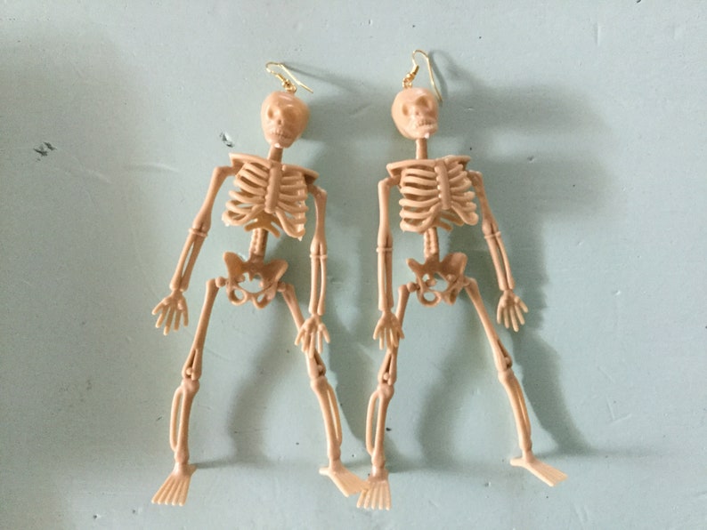 May include: A pair of beige plastic skeleton earrings. Each earring features a miniature skeleton with articulated limbs and a gold-colored hook for the ear. The earrings are designed to dangle and are set against a light blue background.