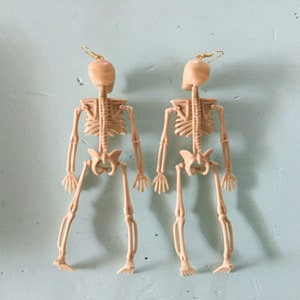 May include: Two beige skeleton earrings with gold-colored ear wires. The earrings are detailed with the skeletal structure of the human body. The earrings are a unique accessory for Halloween or gothic-themed outfits.