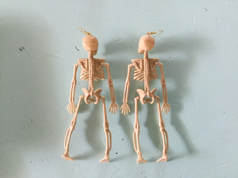 May include: Two beige skeleton earrings with gold-colored ear wires. The earrings are detailed with the skeletal structure of the human body. The earrings are a unique accessory for Halloween or gothic-themed outfits.