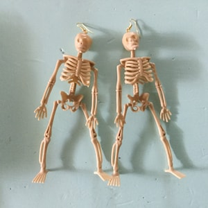 May include: A pair of beige plastic skeleton earrings with gold-colored ear wires. The earrings are in the shape of a human skeleton, with detailed rib cages, arms, and legs. The earrings are displayed against a light blue background.