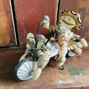 May include: A whimsical sculpture of a motorcycle and rider, crafted entirely from seashells. The rider wears a hat and the motorcycle has a black and white wheel. The word "Florida" is visible on the side. The sculpture is set against a wooden background.