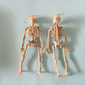 May include: Two beige skeleton earrings with gold-colored ear wires. The earrings are detailed with rib cages, spines, and limbs. The earrings are a fun and quirky accessory, perfect for Halloween or adding a unique touch to any outfit.
