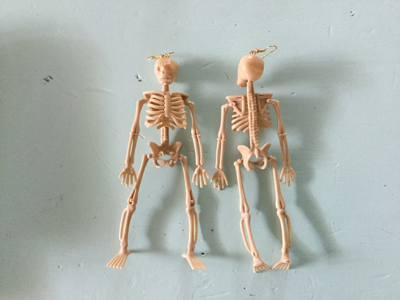 May include: Two beige skeleton earrings with gold-colored ear wires. The earrings are detailed with rib cages, spines, and limbs. The earrings are a fun and quirky accessory, perfect for Halloween or adding a unique touch to any outfit.