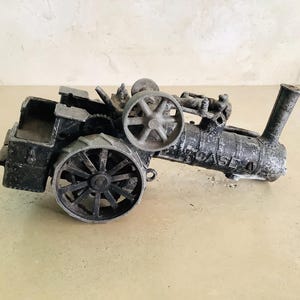May include: A vintage toy tractor made of metal, painted in a weathered gray and black finish. The tractor has a large rear wheel and a smaller front wheel, and features a detailed engine and a pipe.