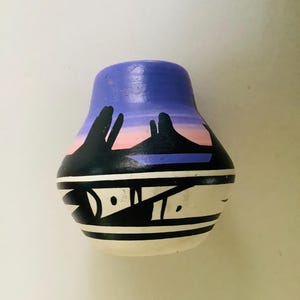 May include: A small, rounded ceramic vase featuring a painted landscape scene with a purple and pink sky above black mountain silhouettes. The lower section of the vase is decorated with a white band containing black geometric shapes and lines.