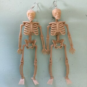 May include: A pair of beige skeleton earrings. Each earring features a miniature skeleton with articulated limbs and a skull head. The earrings hang from silver-colored fish hook ear wires. The skeletons are holding hands.