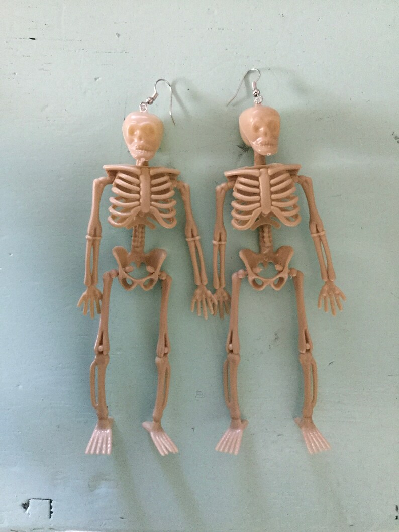 May include: A pair of beige skeleton earrings. Each earring features a miniature skeleton with articulated limbs and a skull head. The earrings hang from silver-colored fish hook ear wires. The skeletons are holding hands.