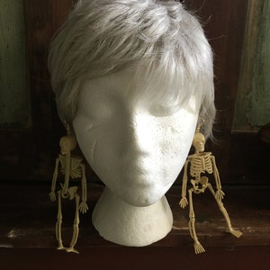 May include: A pair of skeleton earrings. Each earring features a small, beige plastic skeleton figure hanging from a gold-colored hook. The earrings are displayed on a white mannequin head with gray hair.