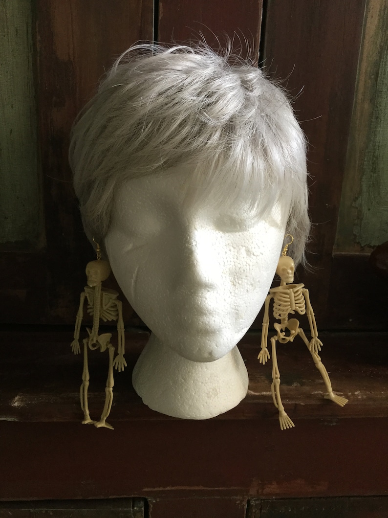 May include: A pair of skeleton earrings. Each earring features a small, beige plastic skeleton figure hanging from a gold-colored hook. The earrings are displayed on a white mannequin head with gray hair.