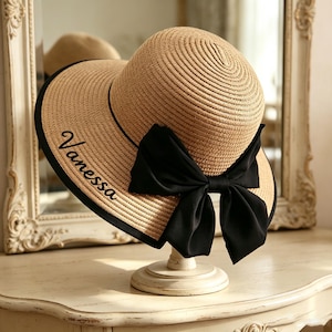 May include: A tan straw sun hat featuring a wide brim with a black fabric trim along the edge. The hat is adorned with a large black fabric bow and the name Vanessa embroidered in black script lettering on the left side of the brim.