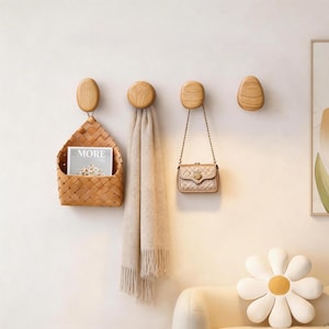 Peut inclure: Quatre patères en bois clair fixées au mur en ligne horizontale. La première patère soutient un panier tressé contenant un magazine avec le texte MORE. La deuxième patère porte une écharpe beige. La troisième porte un petit sac à main doré.