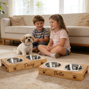 Könnte beinhalten: Zwei Hundenäpfe aus Holz mit Edelstahleinsätzen. Eine Schüssel ist mit "Daisy" und die andere mit "Bella" beschriftet. Beide haben Pfotenabdruck-Designs. Ein kleiner Hund sitzt in der Nähe der Schalen, mit zwei Kindern im Hintergrund.