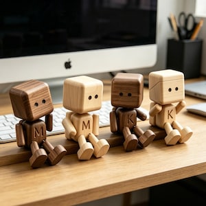 May include: Four wooden block figures with movable limbs and cube-shaped heads. Each figure has a different wood tone, with the letters "M" and "K" carved on the torso. The figures are seated on a wooden desk.