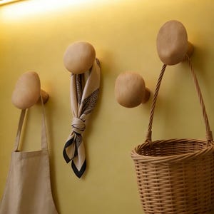 May include: Wooden wall hooks in various shapes, holding a beige apron, a patterned scarf, and a woven basket. The hooks are mounted on a yellow wall, creating a functional and decorative storage solution.