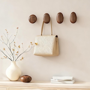 Peut inclure: Quatre patères murales en bois, dont une ronde et trois ovales, avec un sac à main crème suspendu à l'une d'elles. Un vase avec des branches et un petit bol en bois sont posés sur une surface en bois.