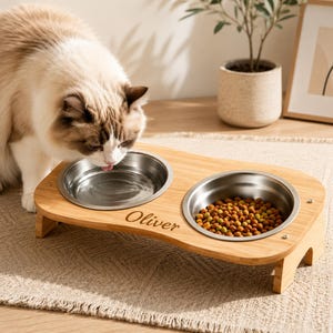 May include: A light brown bamboo elevated pet feeder with two circular stainless steel bowls. The wooden stand features the name Oliver engraved on the front. One bowl contains water and the other contains dry brown kibble for a pet.