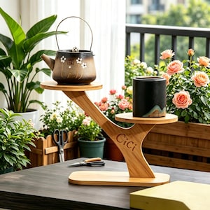 Puede incluir: Un soporte para plantas de bambú con dos estantes circulares, que sostiene una tetera de cerámica marrón y una maceta de cerámica negra. El soporte tiene un diseño único en forma de árbol con la palabra "Cici" grabada en la parte delantera. El fondo presenta plantas y flores.