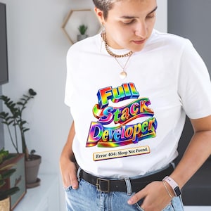 Könnte beinhalten: Weißes T-Shirt mit einem farbenfrohen Grafikdesign mit der Aufschrift "Full Stack Developer". Das Design hat einen 3D-Effekt mit Regenbogenfarben. Darunter steht "Error 404: Sleep Not Found".