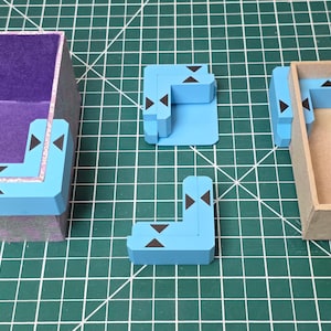 May include: A light blue corner piece with black arrow details is placed near a purple-lined box and a cardboard box. Several other blue corner pieces are scattered on a green grid surface. The pieces appear to be components of a larger structure.