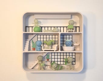 May include: A white multi-level display shelf featuring various small, stylized figurines in different poses. The shelf has three levels with stairs, grid-patterned backdrops, and miniature furniture including a sofa, bathtub, and sink.