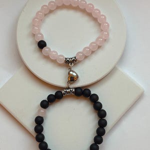 May include: Two beaded bracelets. One is light pink with a silver heart charm, the other is black. The bracelets are connected by a silver heart-shaped magnetic clasp. The bracelets are on a white surface.