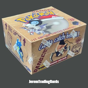 Puede incluir: Una caja de sobres de cartas coleccionables Pokémon Fossil sellada en cartón. La caja de color tostado presenta el logotipo de Pokémon, el texto "Gotta catch 'em all!", "1st Edition Limited Printing" y "Danger!". Muestra ilustraciones de Aerodactyl, Lapras y Kabutops.