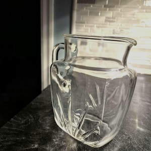 May include: A clear glass pitcher with a square base and a handle. The pitcher has a slightly flared top and a spout for easy pouring. The glass has a textured, faceted design.