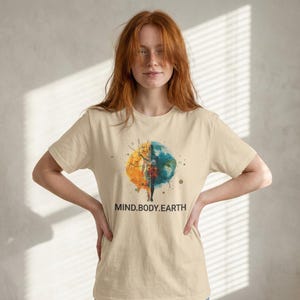 May include: Beige t-shirt featuring a watercolor design of a split globe, half yellow and orange, half blue and green, with the text "MIND.BODY.EARTH" below. The shirt has short sleeves and a crew neck.