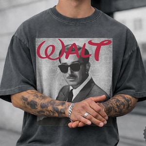 May include: A charcoal gray short-sleeve t-shirt featuring a graphic print of a man in a suit and sunglasses. The text WALT is printed in red above the portrait. The shirt has a faded, washed-out appearance and a crew neckline with short sleeves.