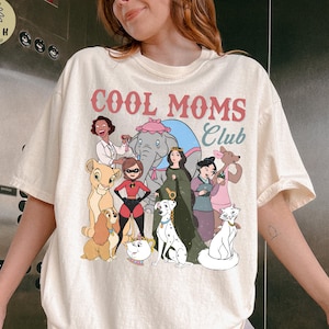 May include: Cream-colored t-shirt with the text "COOL MOMS CLUB" in pink and blue. The shirt features a graphic of various cartoon characters. The shirt is a relaxed fit.
