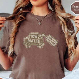 May include: Espresso-colored t-shirt featuring a distressed graphic of a tow truck with the words "Tow Mater" and "Towing & Salvage." The shirt is a comfort color and has a relaxed fit. The shirt is a brown color.