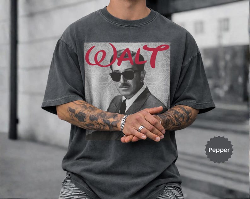 May include: A charcoal gray short-sleeve t-shirt featuring a graphic print of a man in a suit and sunglasses. The text WALT is printed in red above the portrait. The shirt has a faded, washed-out appearance and a crew neckline with short sleeves.