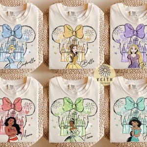 Könnte beinhalten: Sechs cremefarbene T-Shirts mit Disney-Prinzessinnen-Designs. Jedes Shirt hat eine große Schleife und eine Prinzessinnen-Illustration innerhalb einer Mickey-Mouse-Kopf-Umrandung. Die Prinzessinnen sind Cinderella, Belle, Rapunzel, Moana, Tiana und Jasmine.