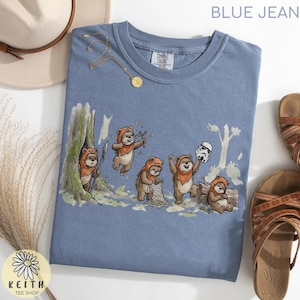 May include: Blue jean colored t-shirt featuring a whimsical illustration of Ewoks in a forest setting. The design includes Ewoks with spears, slingshots, and a Stormtrooper helmet. The shirt has a Comfort Colors tag and the text "BLUE JEAN" at the top.