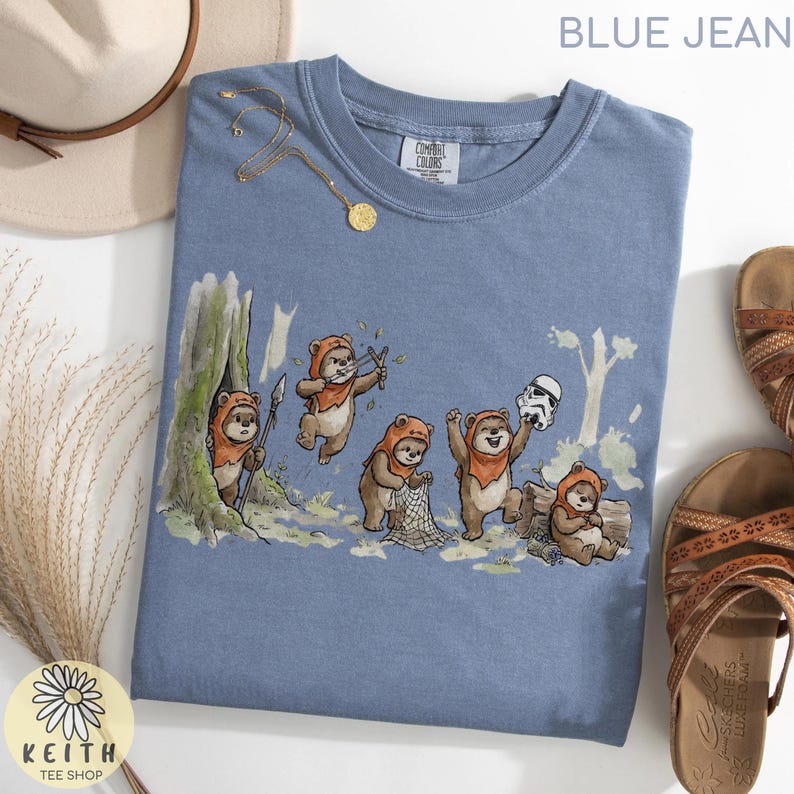 May include: Blue jean colored t-shirt featuring a whimsical illustration of Ewoks in a forest setting. The design includes Ewoks with spears, slingshots, and a Stormtrooper helmet. The shirt has a Comfort Colors tag and the text "BLUE JEAN" at the top.