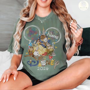 May include: Green t-shirt featuring a Disney-themed graphic with the text "Disney 2020". The design includes characters from Beauty and the Beast, fireworks, and a castle. The word "Name" is also present in the design.