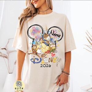 May include: Cream-colored t-shirt featuring a Disney-themed graphic. The design includes characters from Beauty and the Beast, fireworks, and the text "Disney 2026". The shirt also has a Mickey Mouse ear design with the word "Name" in it.
