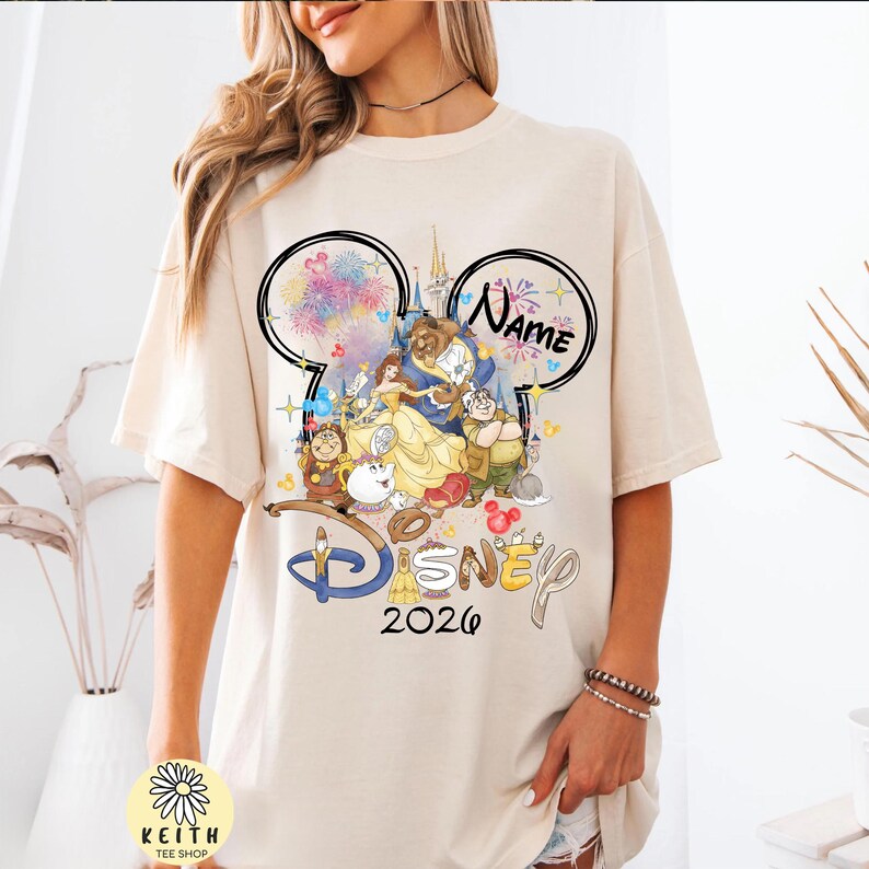May include: Cream-colored t-shirt featuring a Disney-themed graphic. The design includes characters from Beauty and the Beast, fireworks, and the text "Disney 2026". The shirt also has a Mickey Mouse ear design with the word "Name" in it.