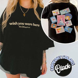 May include: Black t-shirt with the text "wish you were here" and "Walt Disney World" in a cream color. The back of the shirt features several colorful postage stamp-like patches with Disney-themed designs. The shirt is part of the Comfort Colors collection.
