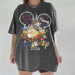 May include: Dark gray t-shirt featuring a Disney-themed graphic. The design includes the text "Name" and "2026" along with illustrations of characters from Beauty and the Beast. The shirt has short sleeves and a relaxed fit.