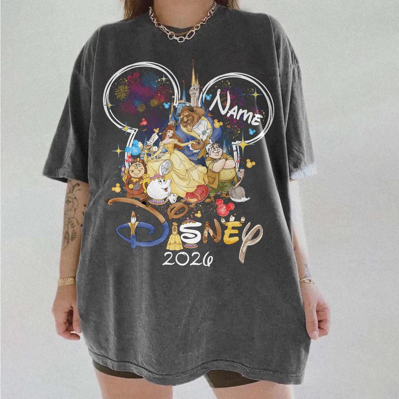 May include: Dark gray t-shirt featuring a Disney-themed graphic. The design includes the text "Name" and "2026" along with illustrations of characters from Beauty and the Beast. The shirt has short sleeves and a relaxed fit.